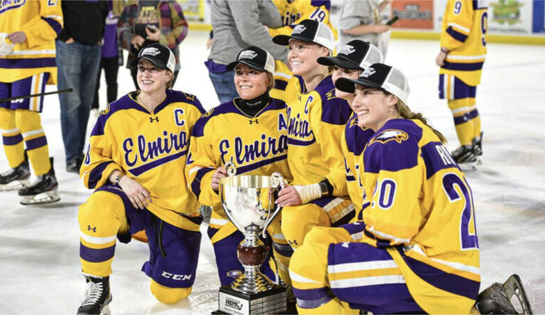 Members of the women's hockey team wear championship hats as they pose on the ice with a trophy