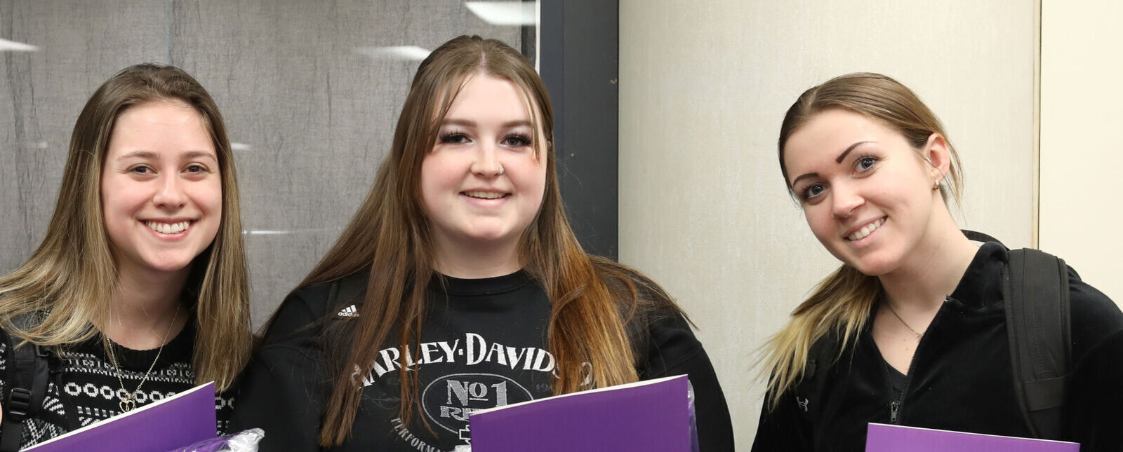 Three female students smile while holding folders.