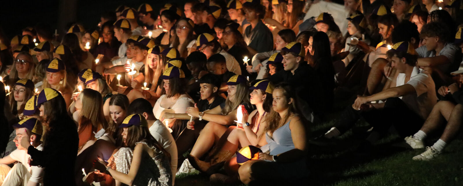 A crowd of students sit on the lawn at night holding lit candles
