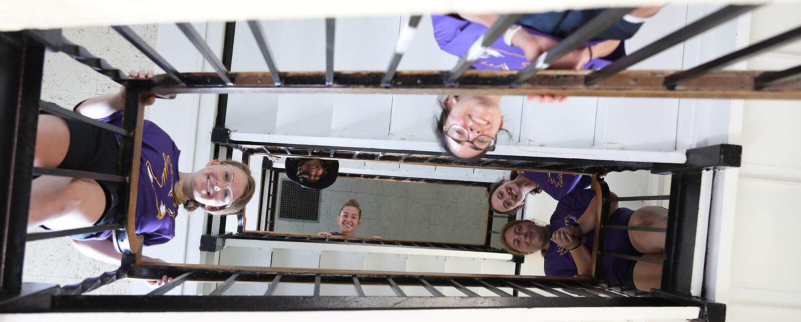 Students look over the railings in a spiraling staircase and smile while looking down at the camera 
