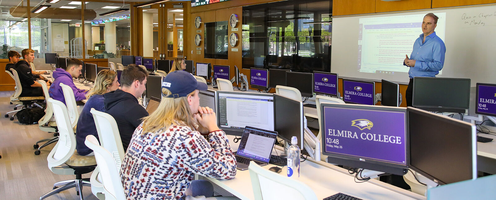 Students work at computers in the Malesardi Finance Trading Room while a professor teaches a finance class