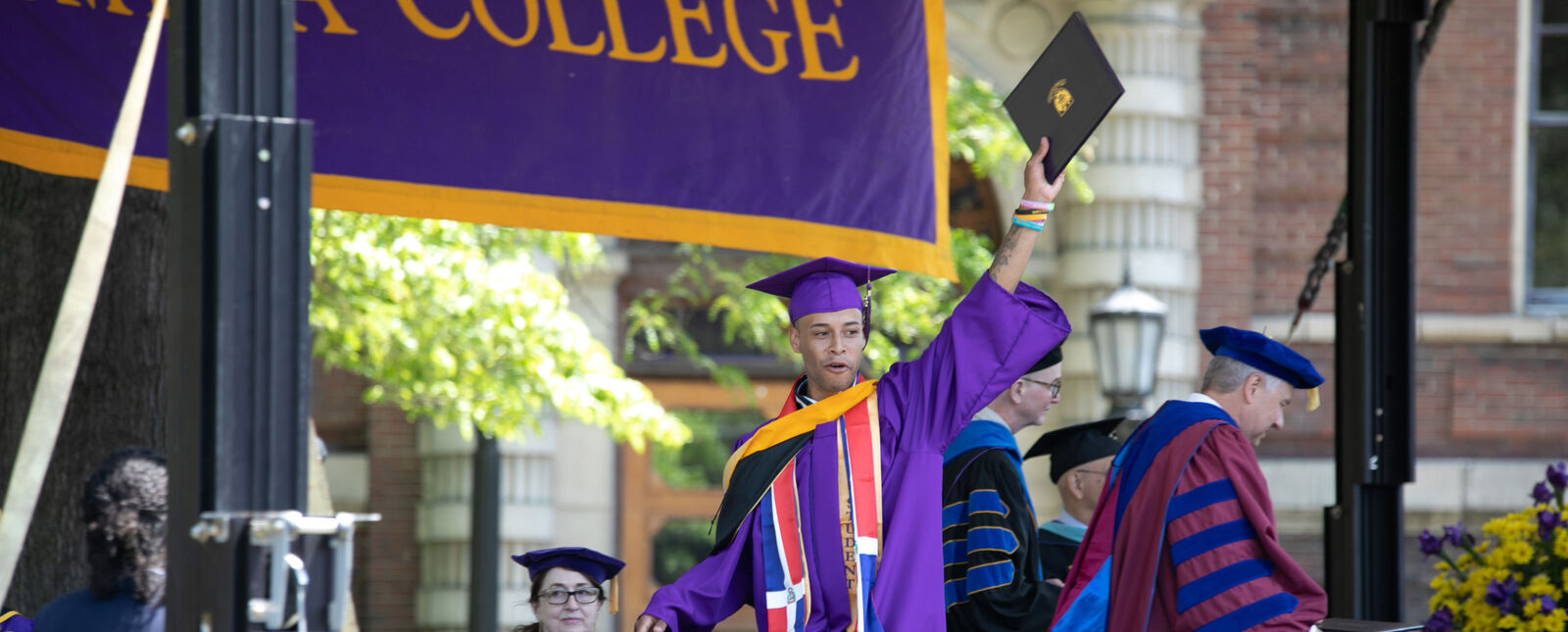 A male graduate raises his degree in celebration from the commencement stage