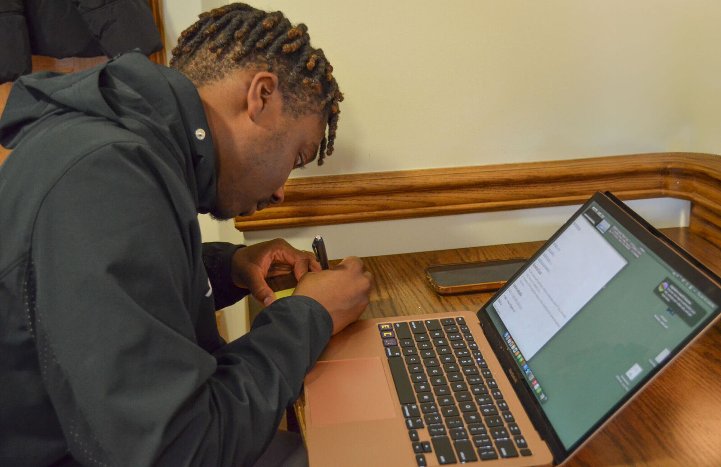 A male student writes a note while working on a laptop on a desk in a dorm room