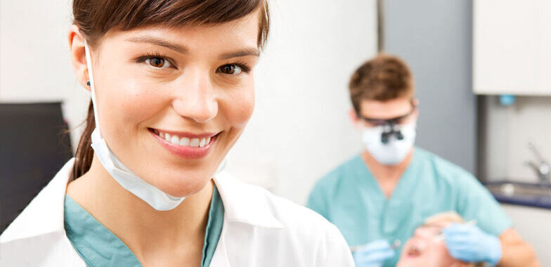 A female dental assistant smiles as a male dental assistant works on a patient in the background