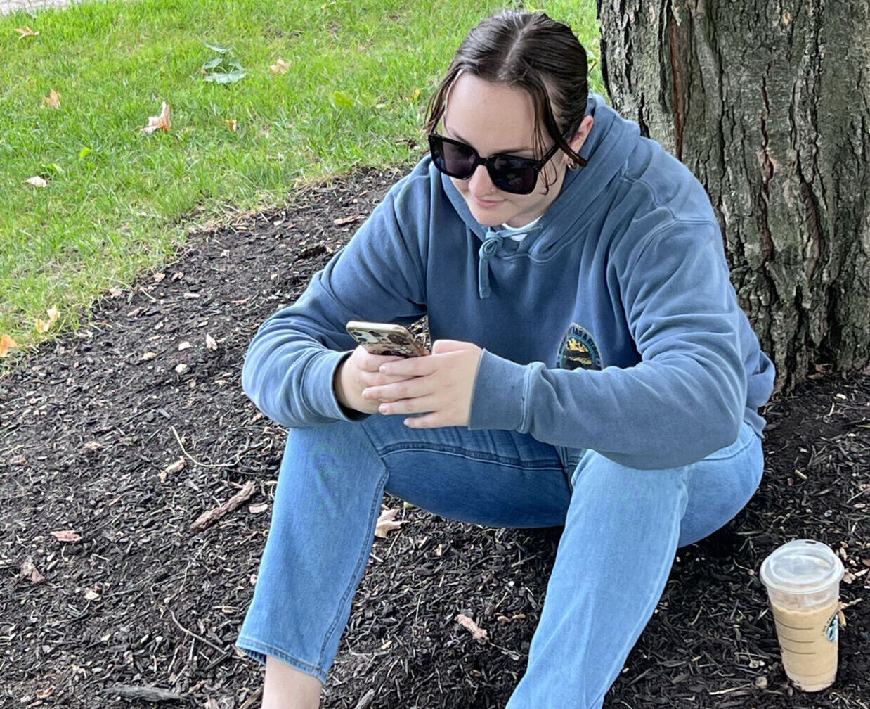 A student checks her phone while sitting under a tree on campus