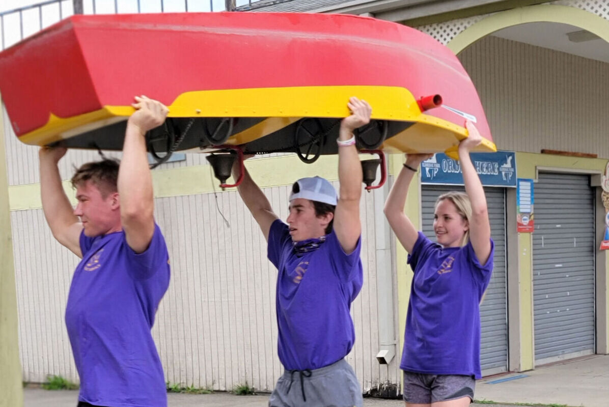 Two male students and a female student carry a boat above their heads across Eldridge Park during a community service outing