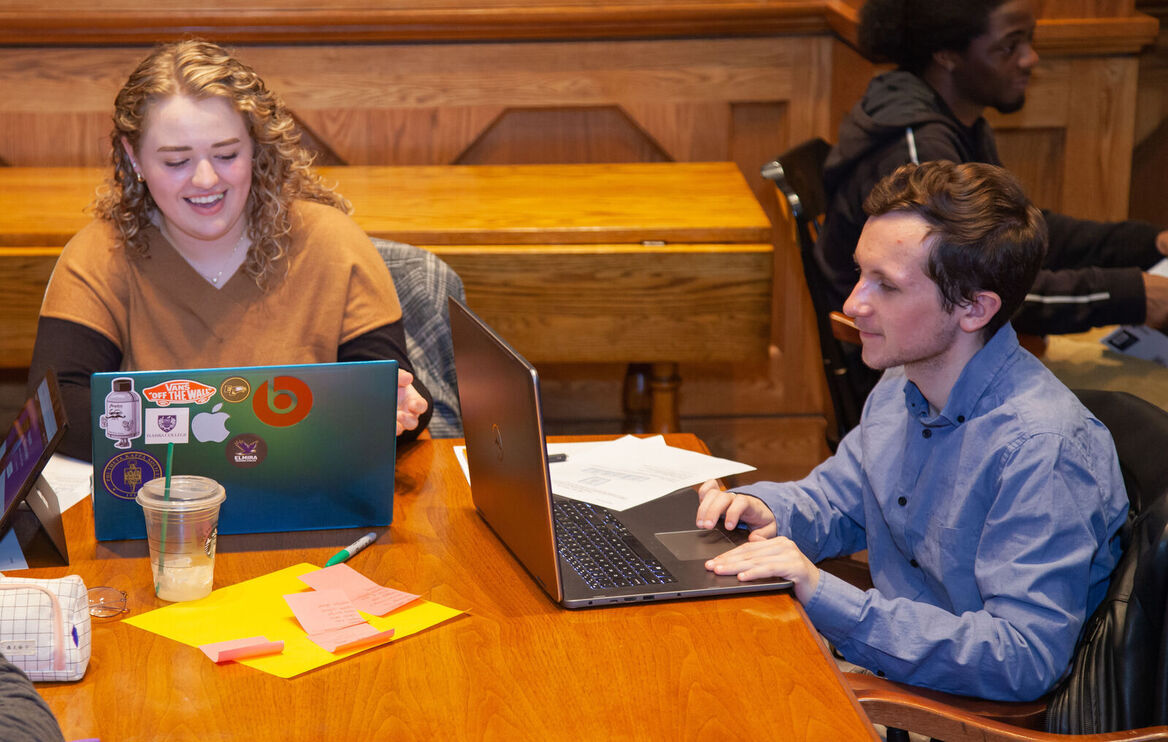 A female student laughs while she works on a laptop at a table near a male student.