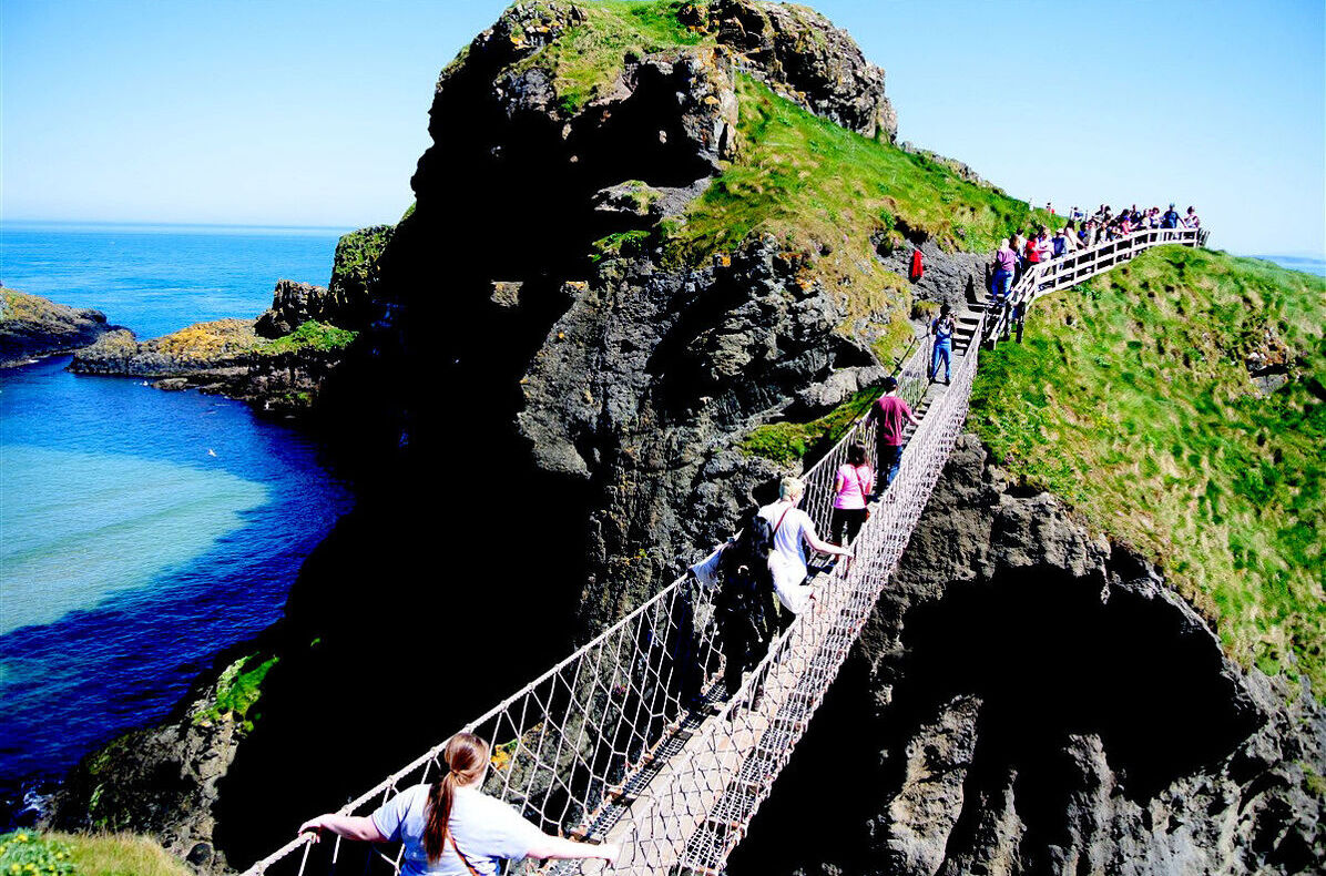 Visitors make their way across a rope bridge spanning a gap in a rocky shore area in Ireland