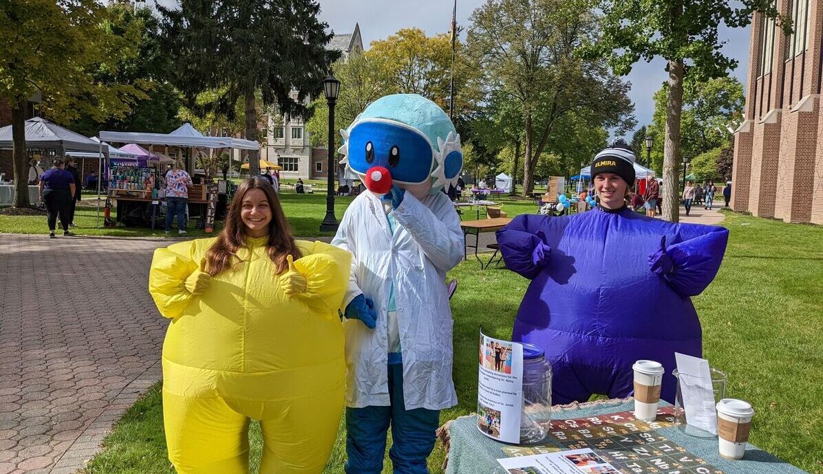 Chemistry Club members wear purple and gold inflatable suits, and stand with a cartoony scientist character during Octagon Fair