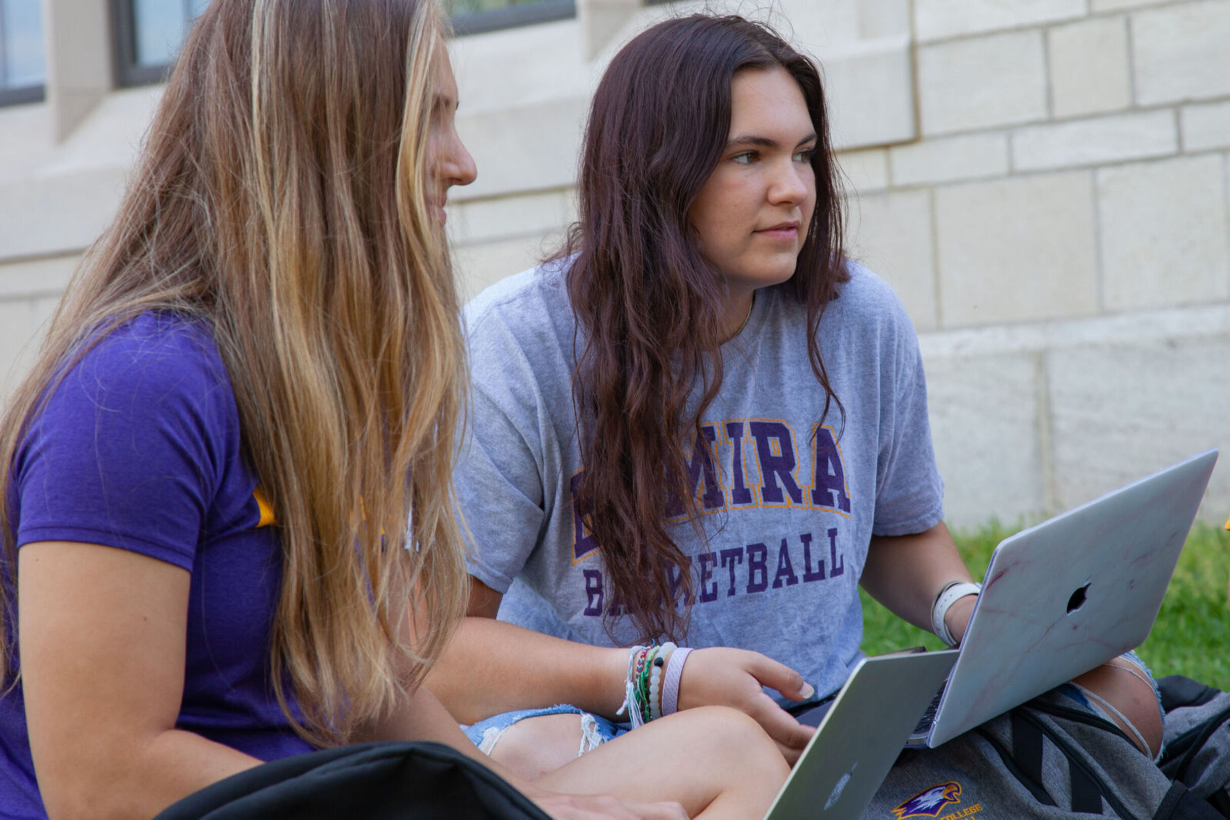 Two female students sit together on the grass while holding laptops on their laps