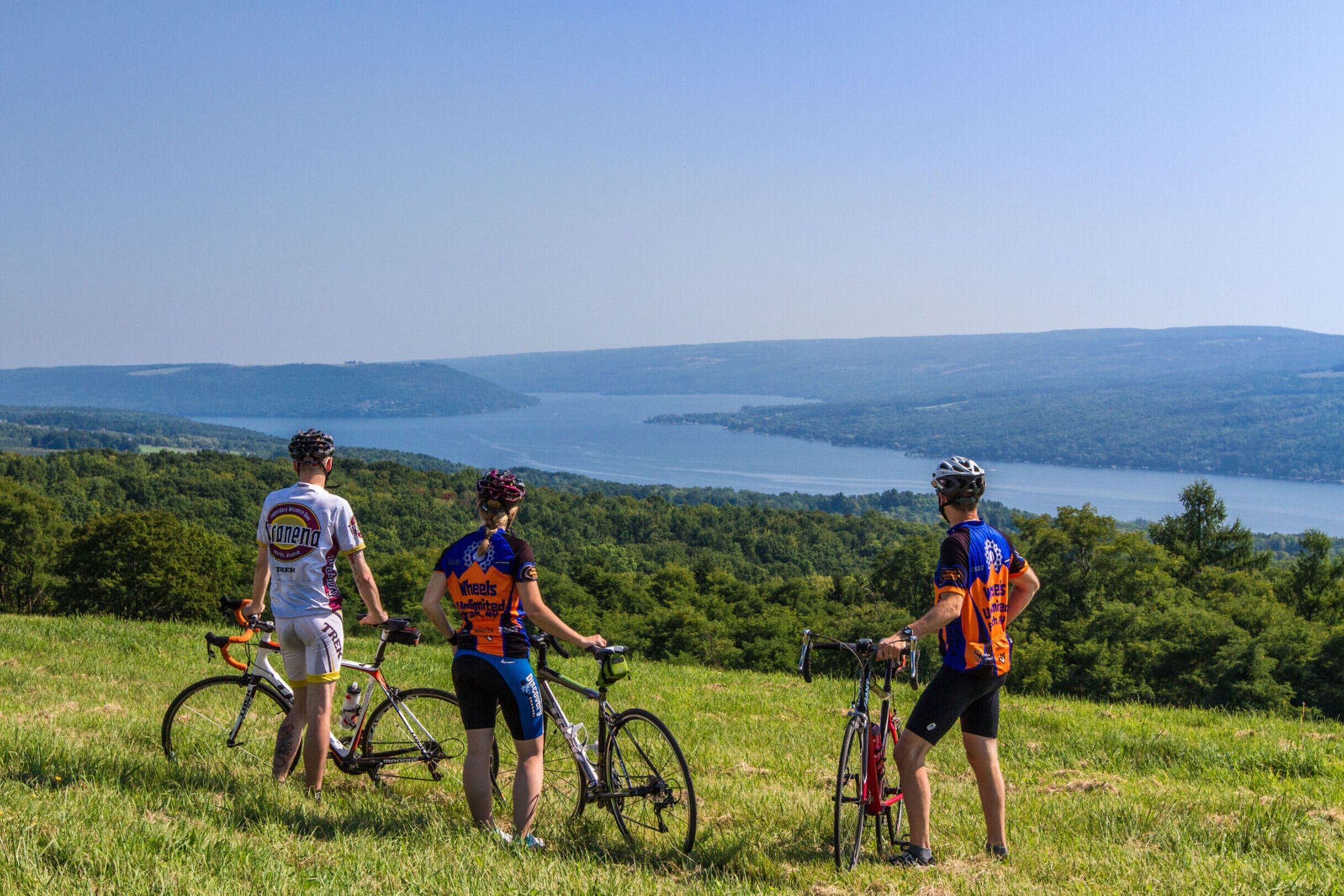 Three cyclists take a break in a a grassy area and look over Keuka Lake