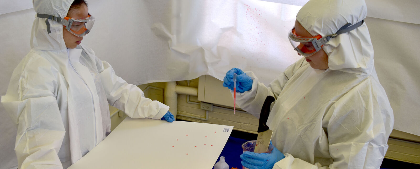 Two students wearing sterile suits see how fake blood can splatter on a surface.