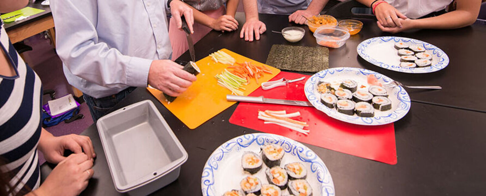 A table with plates of hand-made sushi. There are people standing around the table but you can only see parts of their bodies, like their hands. 
