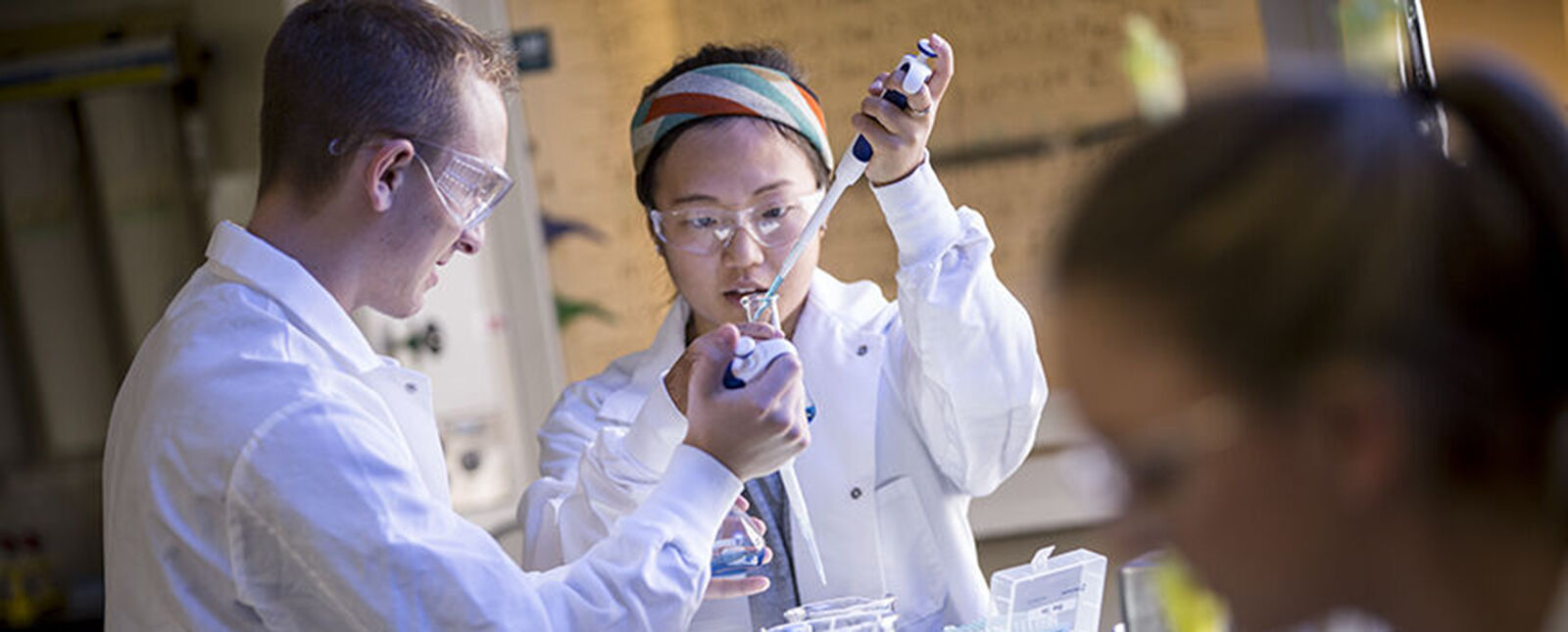A male student holds a glass container as a female student uses a dropper to add a light blue liquid
